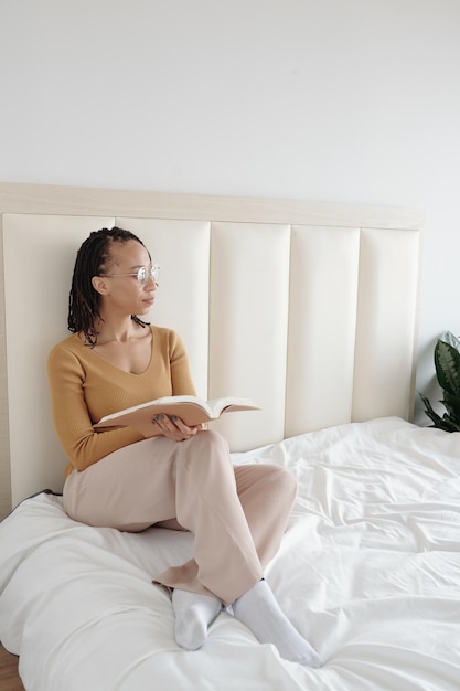 Pensive woman sitting on bed avec livre ouvert et à l'écart