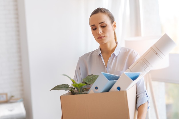 Perdre un emploi. Triste jeune femme calme et frustrée en rentrant à la maison avec une lourde boîte pleine d'objets personnels après avoir perdu son emploi