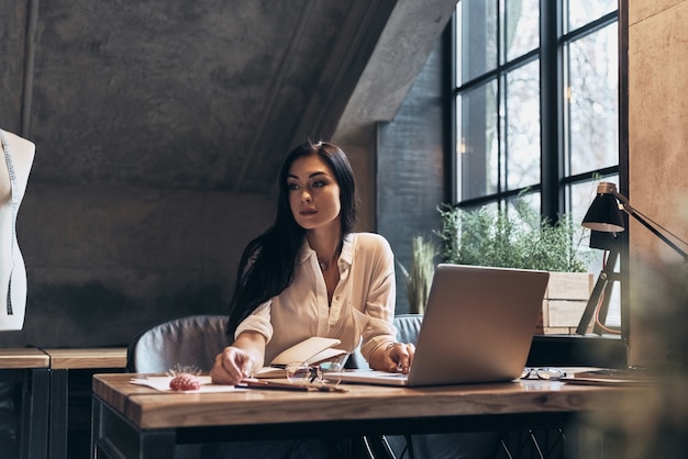 Perdu dans les pensées. Belle jeune femme travaillant à l'aide d'un ordinateur portable et regardant ailleurs alors qu'elle était assise dans son atelier