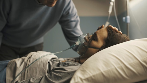 Père âgé et attentionné aidant sa fille allongée dans un lit d'hôpital avec un masque à oxygène, il lui touche la tête