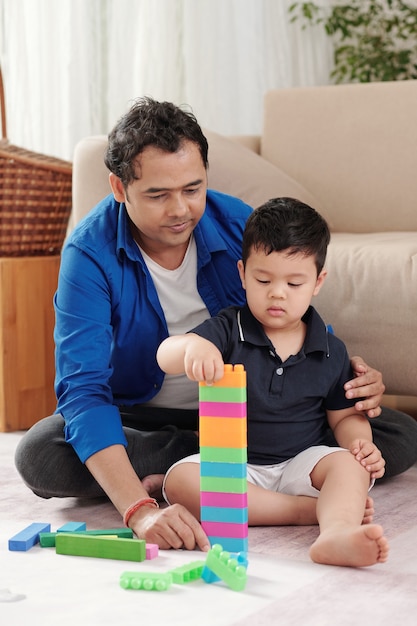 Père aidant son petit fils à construire une haute tour avec des blocs de jouets en plastique colorés
