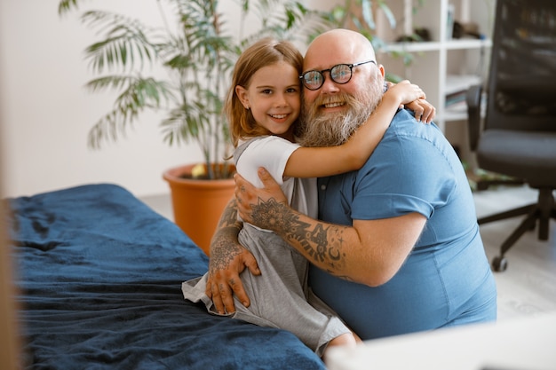 Un père barbu souriant avec des lunettes embrasse une jolie petite fille blonde assise sur le lit à la maison. Soutien affectif en famille