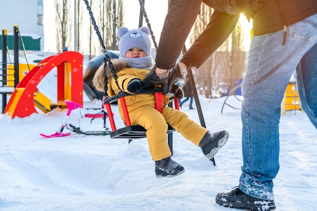 Le père berce un tout-petit sur une balançoire sur une aire de jeux d'hiver