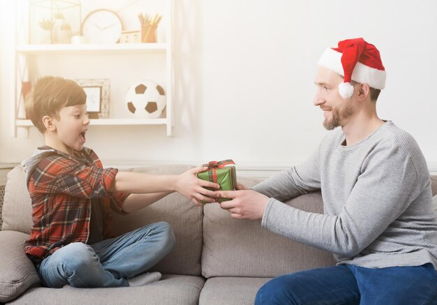 Père en bonnet de Noel donnant à son fils un cadeau de Noël à la maison dans le salon