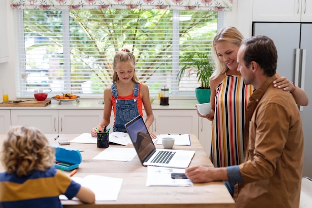 Père caucasien travaillant à la maison sur un ordinateur portable parlant à sa femme, avec son fils et sa fille faisant des travaux scolaires