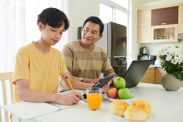 Père contrôlant son fils adolescent étudiant ou faisant ses devoirs à la table de la cuisine à la maison