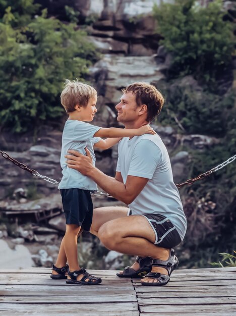 Père étreint son fils assis accroupi se regarde sourire Bonne enfance fête des pères fond de chaîne de montagne comme symbole d'un lien fort entre papa et enfant Influence du père sur la vision du monde de son fils