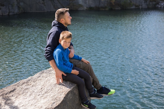 Père de famille heureux jouant avec son fils assis sur un rocher en profitant de la vue sur un lac forestier une belle journée d'automne