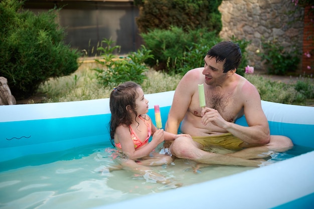 Père et fille aimants profitant du week-end ensemble assis dans une piscine gonflable et mangeant de la glace sucrée