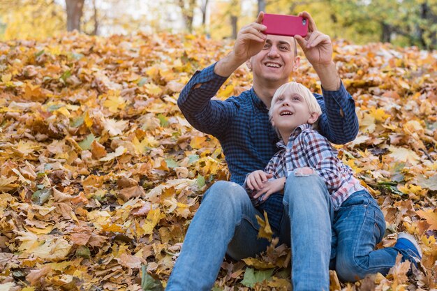 Père et fils assis sur les feuilles d'automne et font des photos au téléphone.