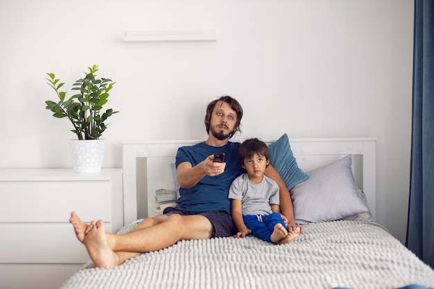 Le père et le fils barbus sont assis sur le lit en T-shirts et regardent un match de football à la télévision. un homme tient une télécommande dans ses mains pour changer de chaîne