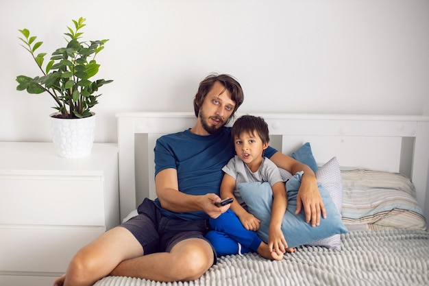 Le père et le fils barbus sont assis sur le lit en T-shirts et regardent un match de football à la télévision. un homme tient une télécommande dans ses mains pour changer de chaîne