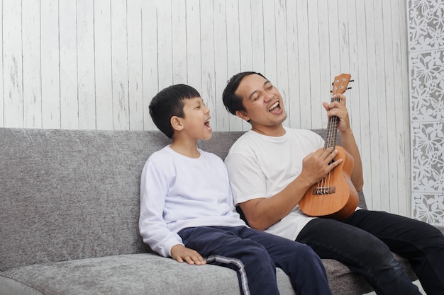 Père et fils chantent ensemble tout en jouant du ukulélé portant un t-shirt blanc assis sur un canapé pl