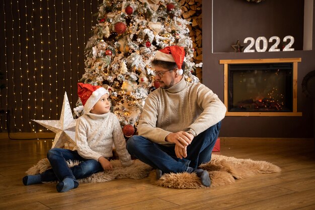 Père et fils en chapeaux de Noël profitent de près de l'arbre de Noël dans une maison à la décoration moderne, heureux nouveau...