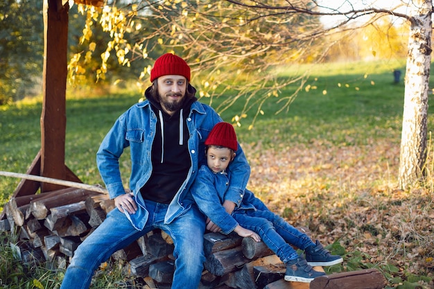 Père et fils en chapeaux rouges assis sur des bûches préparées pour un feu de joie en automne au coucher du soleil