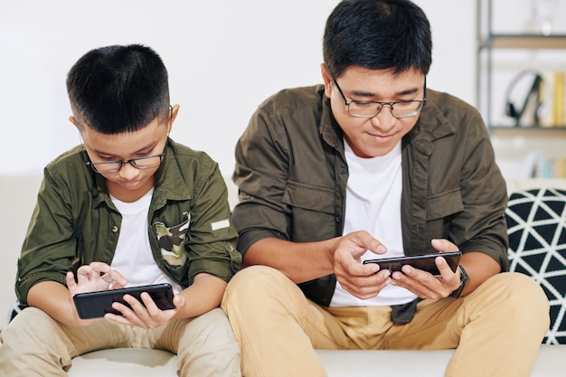 Père et fils vietnamiens dans des verres assis sur un canapé et jouant des jeux sur smartphones