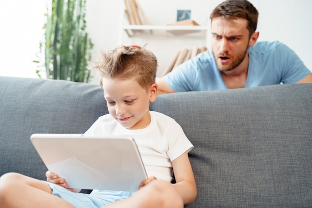 Père inquiet de regarder ce que son fils regarde sur une tablette numérique assis sur un canapé à la maison