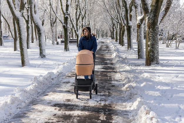 Père marche avec une poussette dans un parc enneigé en hiver. Passer du temps avec bébé dans une belle journée d'hiver. Promenades récréatives avec un enfant. Homme dans une veste anorak bleue.