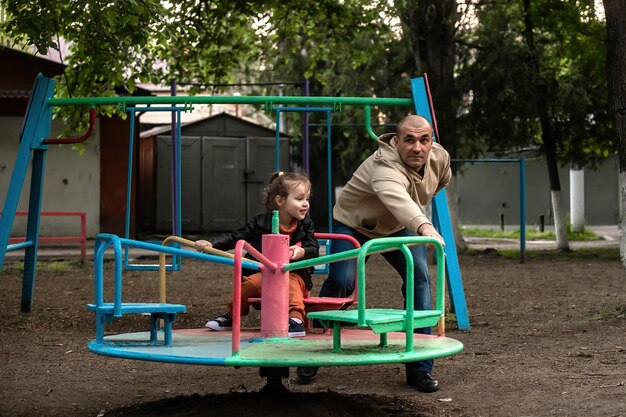 père marche avec sa petite fille, la roule sur une balançoire ronde dans la cour de la maison