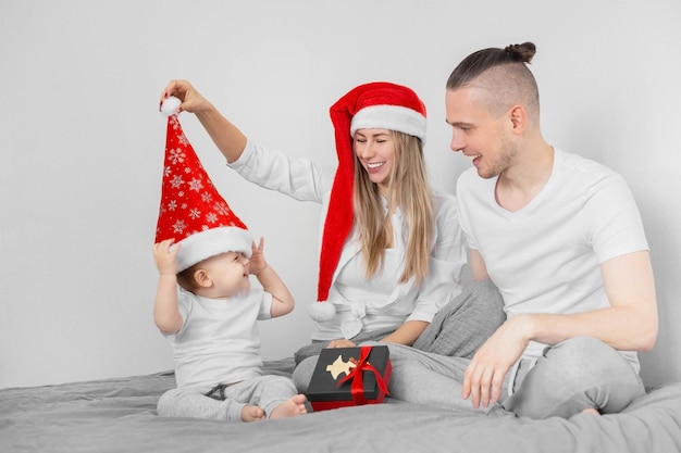 Père de mère et leur fils de bébé dans des chapeaux de Santa s'asseyant sur le lit avec un cadeau de Noël