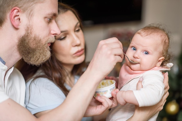 Photo père et mère nourrissent leur bébé de purée de fruits dans la cuisine à partir d'une cuillère