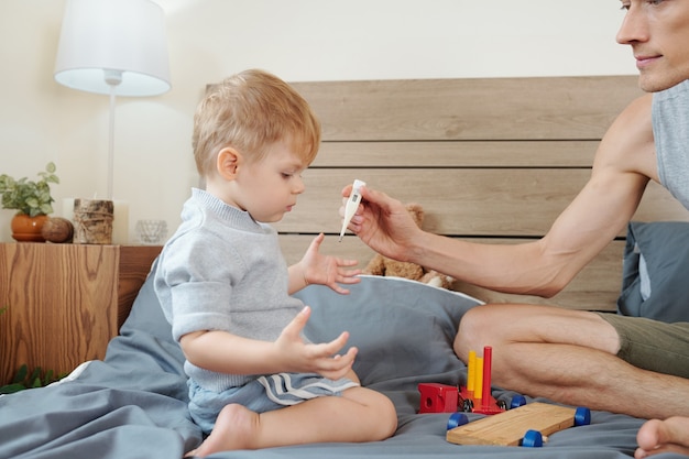 Père mesurant la température de son petit fils pendant qu'ils jouent sur le lit