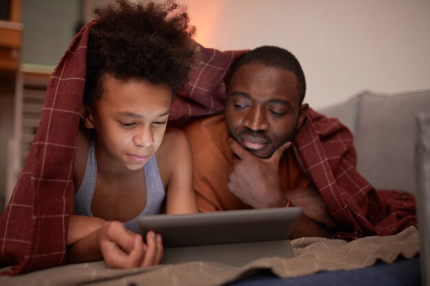 Un père moderne passe la soirée avec son adorable fils à la maison à regarder des dessins animés en ligne sur une tablette numérique