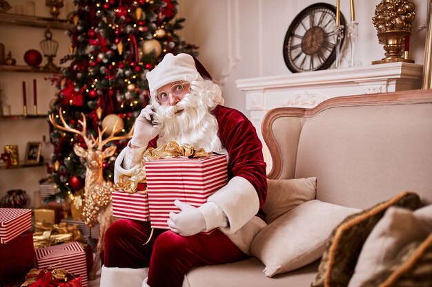 Père Noël assis sur un canapé et parler au téléphone mobile près de la cheminée et de l'arbre de Noël avec des cadeaux. Nouvel an et joyeux Noël, concept de joyeuses fêtes