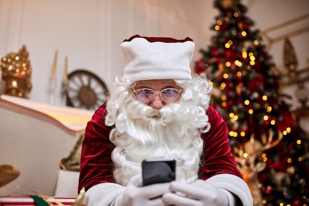 Père Noël assis sur un canapé et parler au téléphone mobile près de la cheminée et de l'arbre de Noël avec des cadeaux.