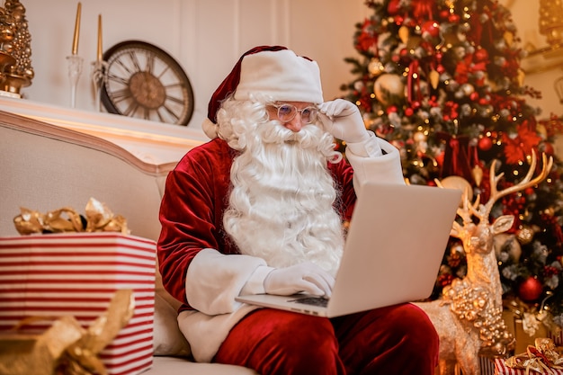 Le Père Noël assis chez lui et lisant des e-mails sur un ordinateur portable avec une demande de hristmas ou une liste de souhaits près de la cheminée et de l'arbre avec des cadeaux. Nouvel an et joyeux Noël, concept de joyeuses fêtes