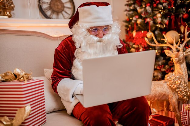 Le Père Noël assis chez lui et lisant des e-mails sur un ordinateur portable avec une demande de Noël ou une liste de souhaits près de la cheminée et un arbre avec des cadeaux. Nouvel an et joyeux Noël, concept de joyeuses fêtes