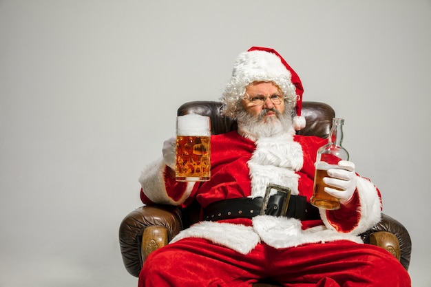 Père Noël buvant de la bière assis sur un fauteuil, félicitant, a l'air ivre et heureux. Modèle masculin caucasien en costume traditionnel. Nouvel An 2020, cadeaux, vacances, ambiance hivernale. Copyspace pour votre annonce.