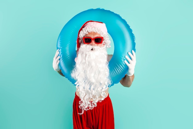 Le Père Noël en costume avec un anneau de natation gonflable dans des lunettes de soleil repose sur la plage sur un fond bleu