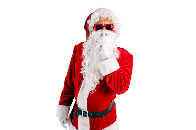 le père Noël en costume rouge avec une barbe dans des lunettes tient le doigt près de ses lèvres et montre un geste de secret