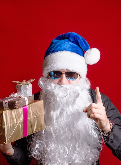 Père Noël dans une casquette bleue sur fond rouge avec des cadeaux secouant son doigt