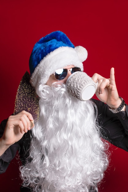 Le Père Noël dans un chapeau bleu sur fond rouge avec des cadeaux boit du lait dans une tasse. Nouvel an Noël