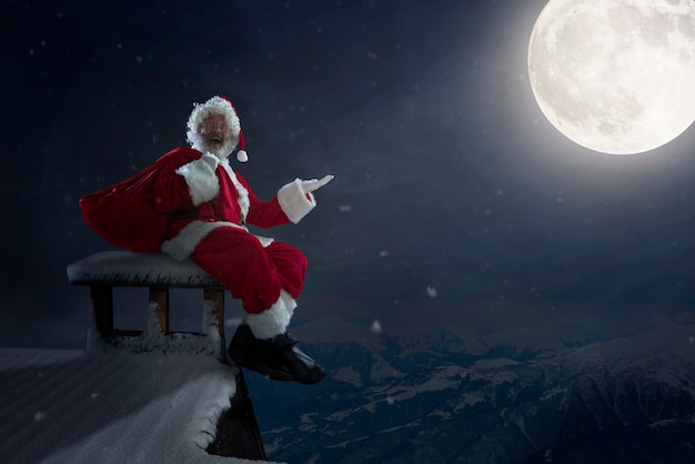 Père Noël émotionnel félicitant pour le Nouvel An 2021 et Noël. Homme en costume traditionnel assis sur le toit de la maison avec la pleine lune sur fond à minuit. Hiver, vacances, soldes. Espace de copie.