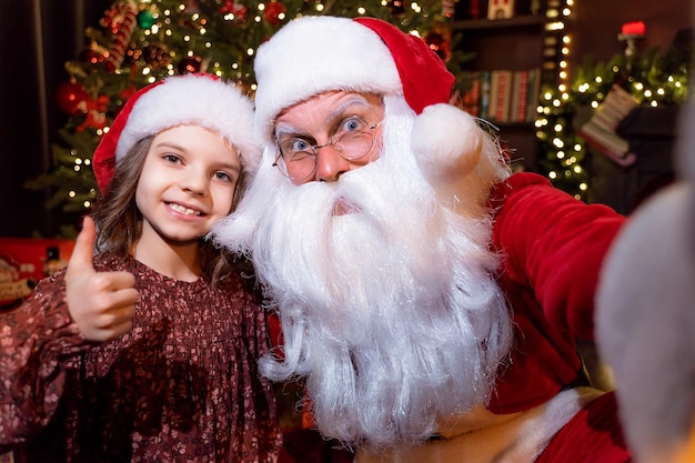 Le Père Noël Fait Un Selfie Avec Une Charmante Petite Fille Qui Montre Le Pouce Vers Le Haut De L'arrière-plan De Noël