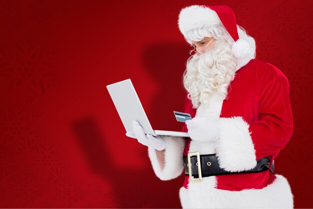 Le père noël fait ses courses en ligne avec un ordinateur portable sur fond de flocon de neige rouge