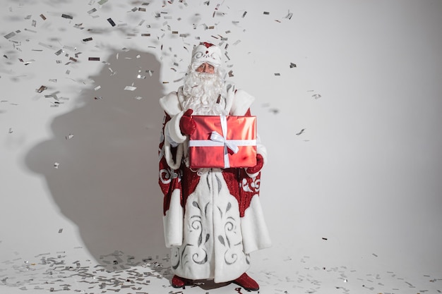 Père Noël avec une longue barbe blanche avec une grande boîte rouge avec un cadeau de Noël dans ses mains, photo isolée sur mur blanc
