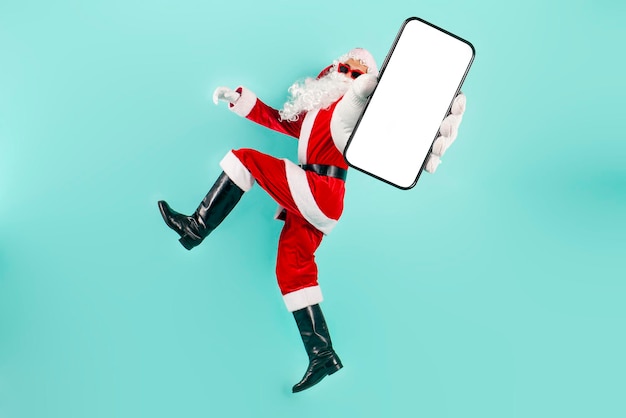 le père Noël en lunettes de soleil et en costume saute et vole et montre un écran de smartphone vierge