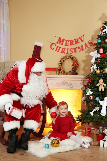 Père Noël offrant un cadeau à une petite fille mignonne près de la cheminée et de l'arbre de Noël à la maison