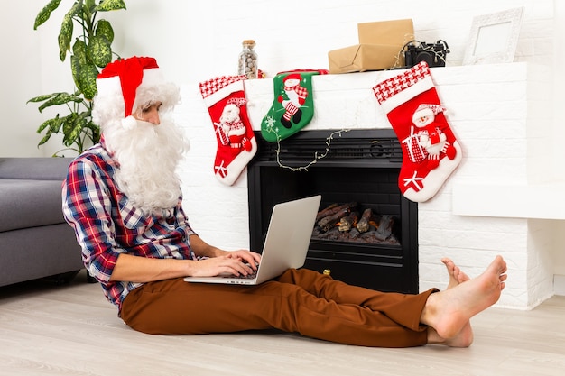 Père Noël avec ordinateur portable assis à table dans la chambre décorée pour Noël