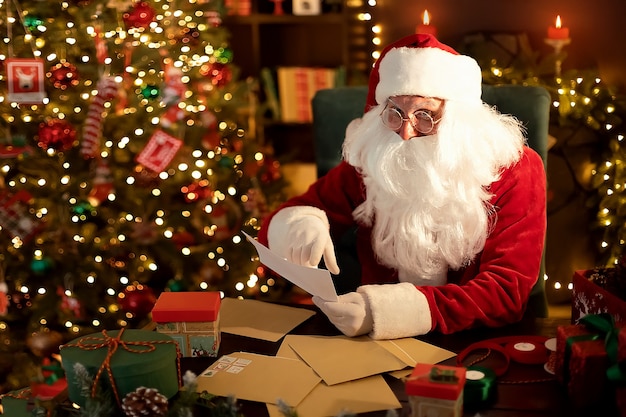 Le père Noël prépare des cadeaux pour les enfants pour Noël à son bureau tout en lisant des listes de souhaits à la maison