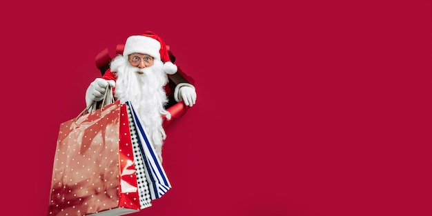 Père Noël regardant à travers le trou sur du papier rouge tout en tenant des sacs à provisions à la main Shopping du Nouvel An