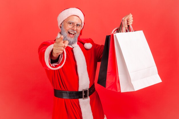 Le père noël regarde la caméra, vous montre du doigt et montre des sacs à provisions, offrant des réductions.