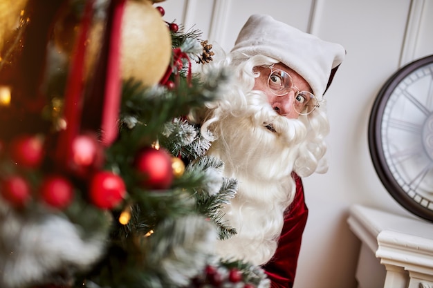 Le Père Noël se cache derrière un arbre de Noël pour cacher des cadeaux.