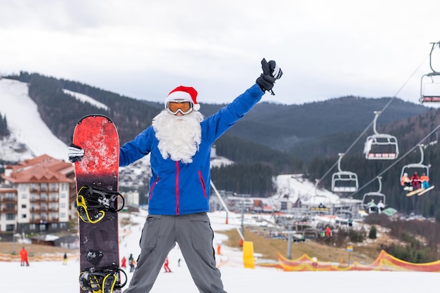 Père Noël avec snowboard contre paysage de station de ski de montagne d'hiver enneigé et ciel bleu, carte de voeux de nouvel an ou de noël.