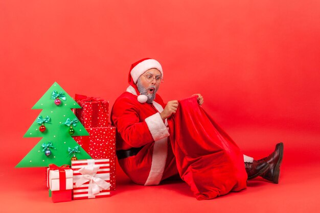 Le père noël surpris assis sur le sol sous un sapin de noël en papier décoré avec des coffrets cadeaux et regardant dans son grand sac rouge avec des cadeaux de vacances. Tourné en studio intérieur isolé sur fond rouge