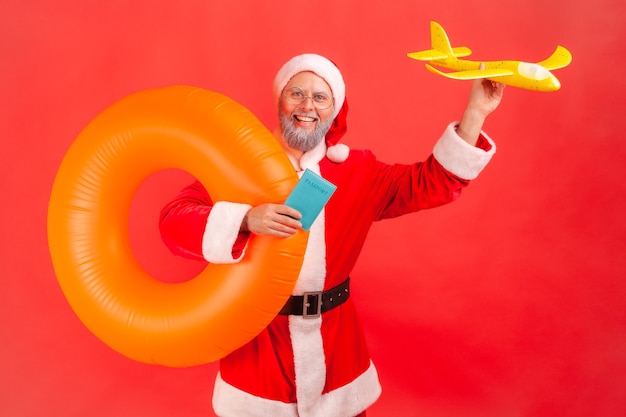 Père noël tenant un anneau en caoutchouc orange, un avion en papier et un passeport, voyageant pendant les vacances d'hiver.